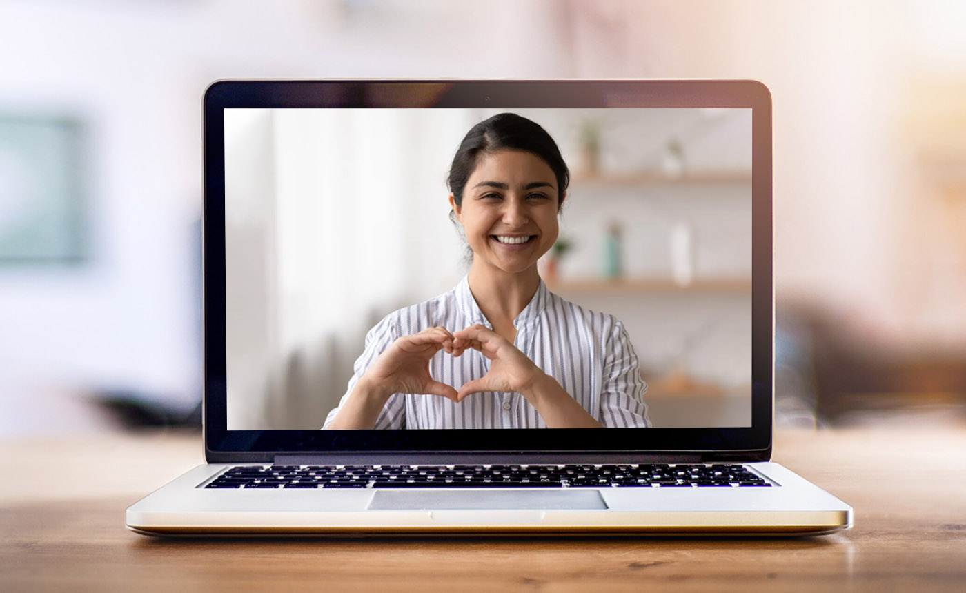 Laptop on a desk with a smiling woman on the screen holding up a heart shape with her hands